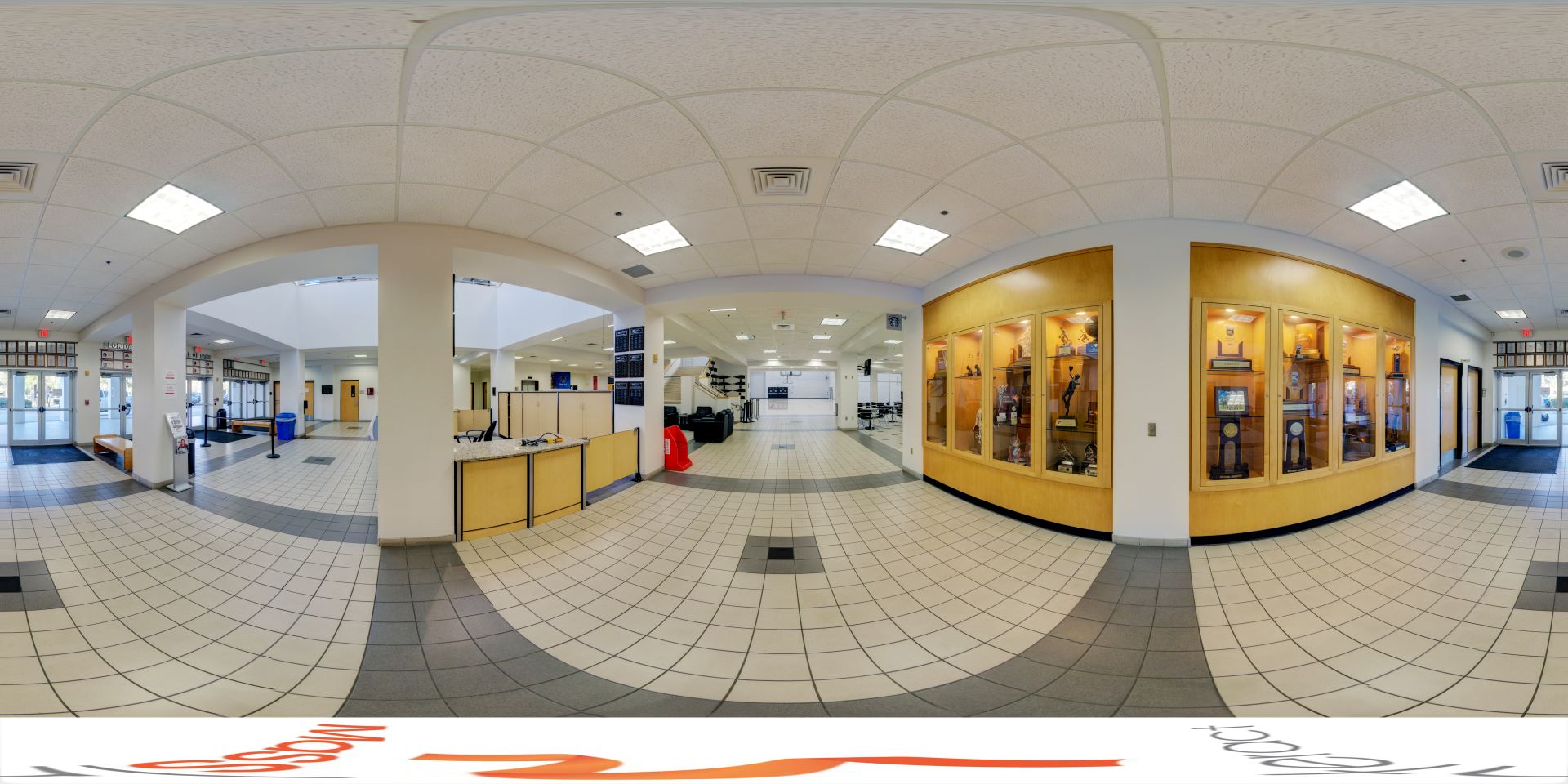 A panoramic view of the Clemente Center lobby at Florida Tech, featuring tiled floors, a reception desk, trophy display cases, seating areas, and multiple entrances with glass doors
